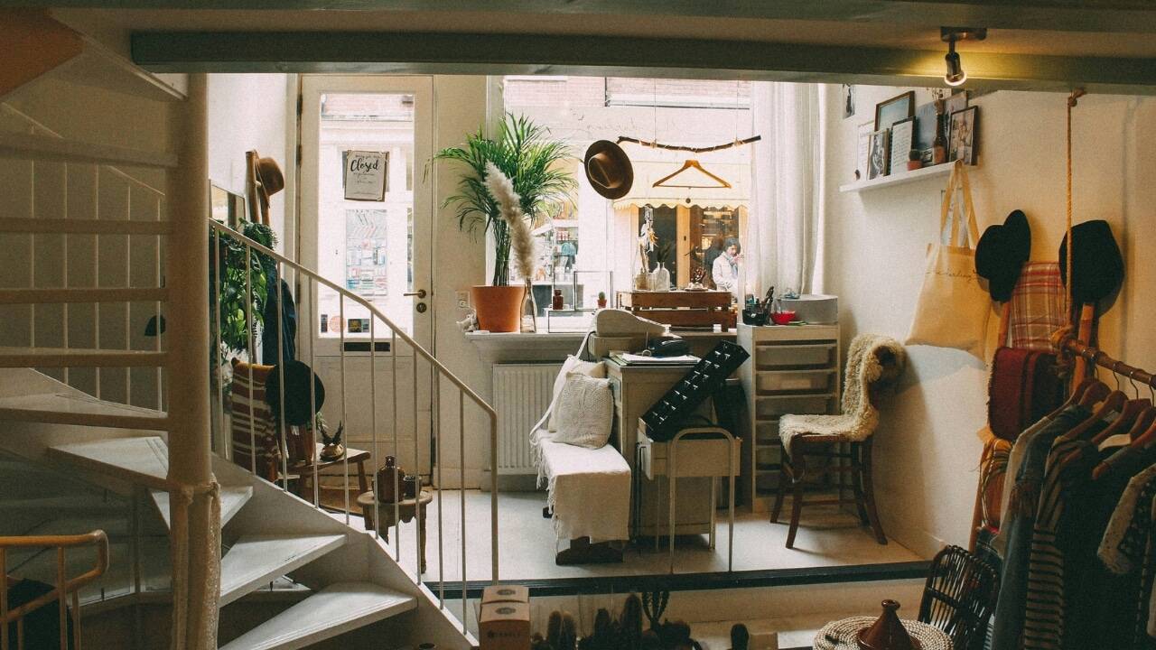 A cluttered room features a white staircase, indoor plants, clothing racks, and various hanging hats.