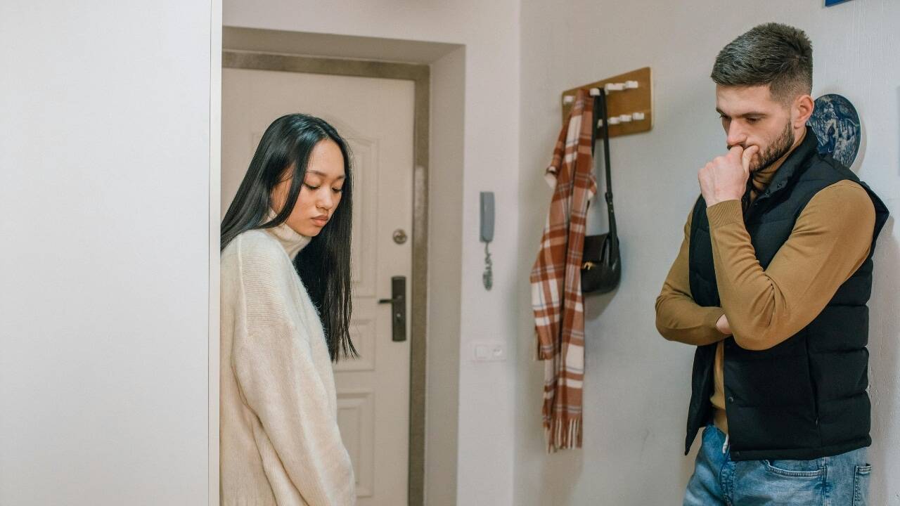 A woman leans against a wall while a man nearby looks down and touches his mouth.