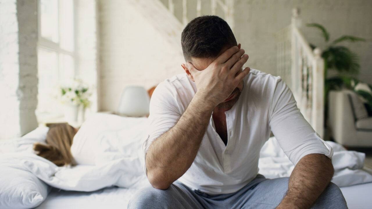 A man sits on a bed covering his face while a woman lies turned away.