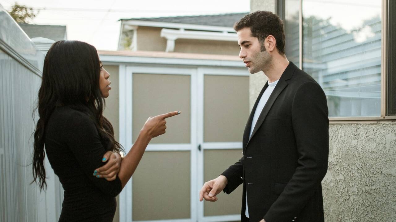 A woman points her finger at a man while they stand outdoors near a white fence.