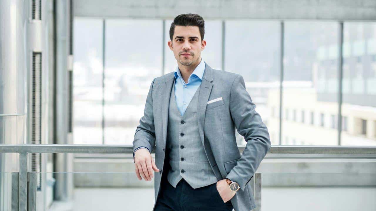 A handsome young businessman in a three-piece suit is standing confidently indoors.